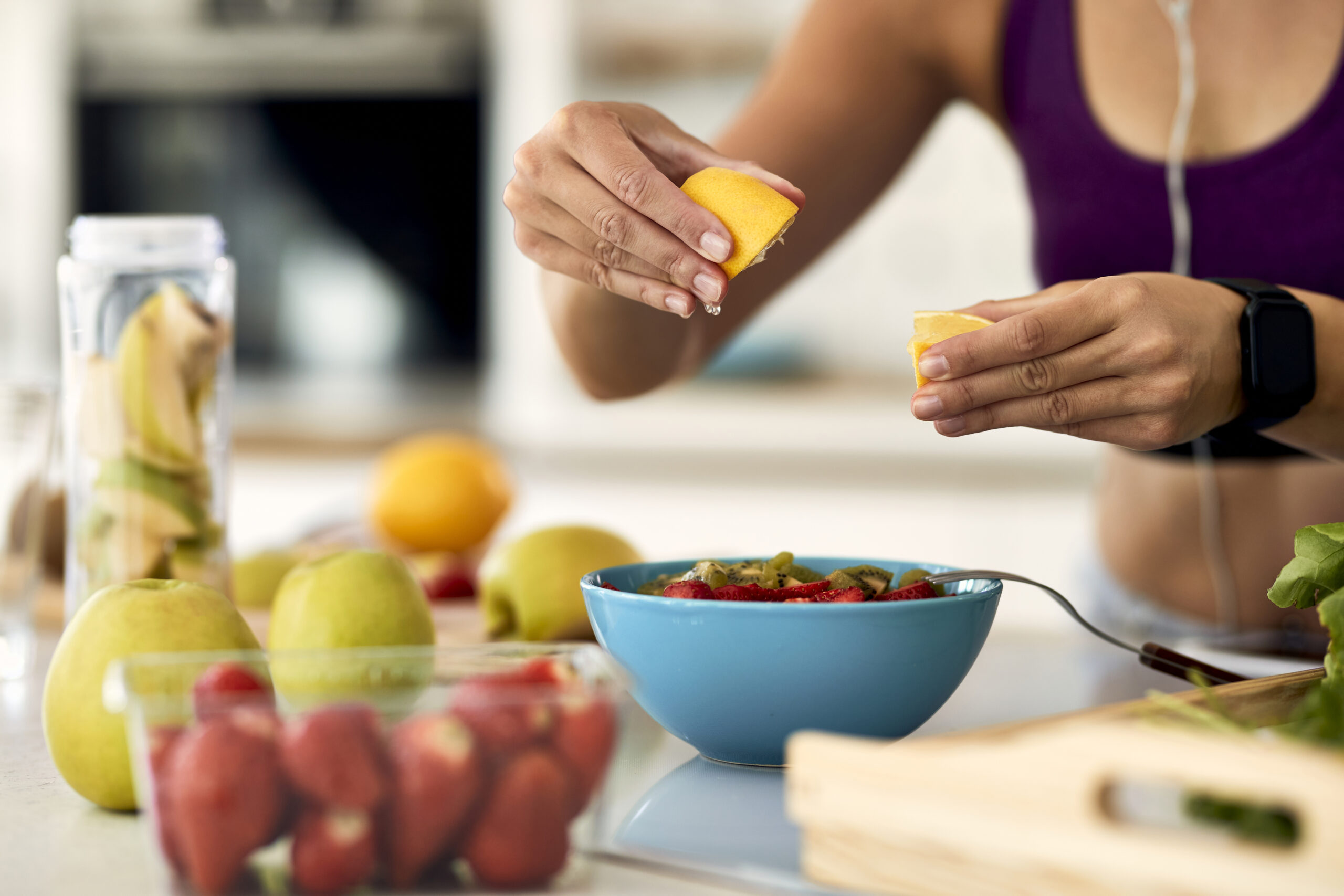 Close-up of athletic woman using lemon while preparing fruit salad in the kitchen.