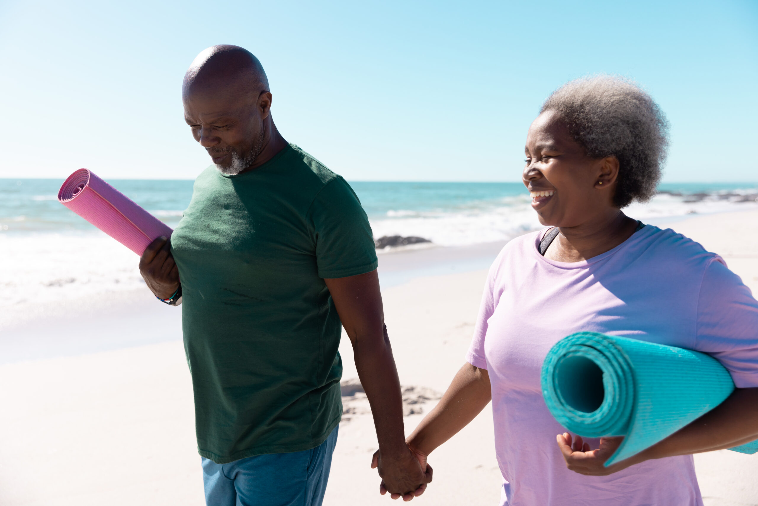 Happy african american senior couple with yoga mats holding hands and walking at beach, copy space. Summer, sky, unaltered, love, together, yoga, retirement, nature, vacation and active lifestyle.
