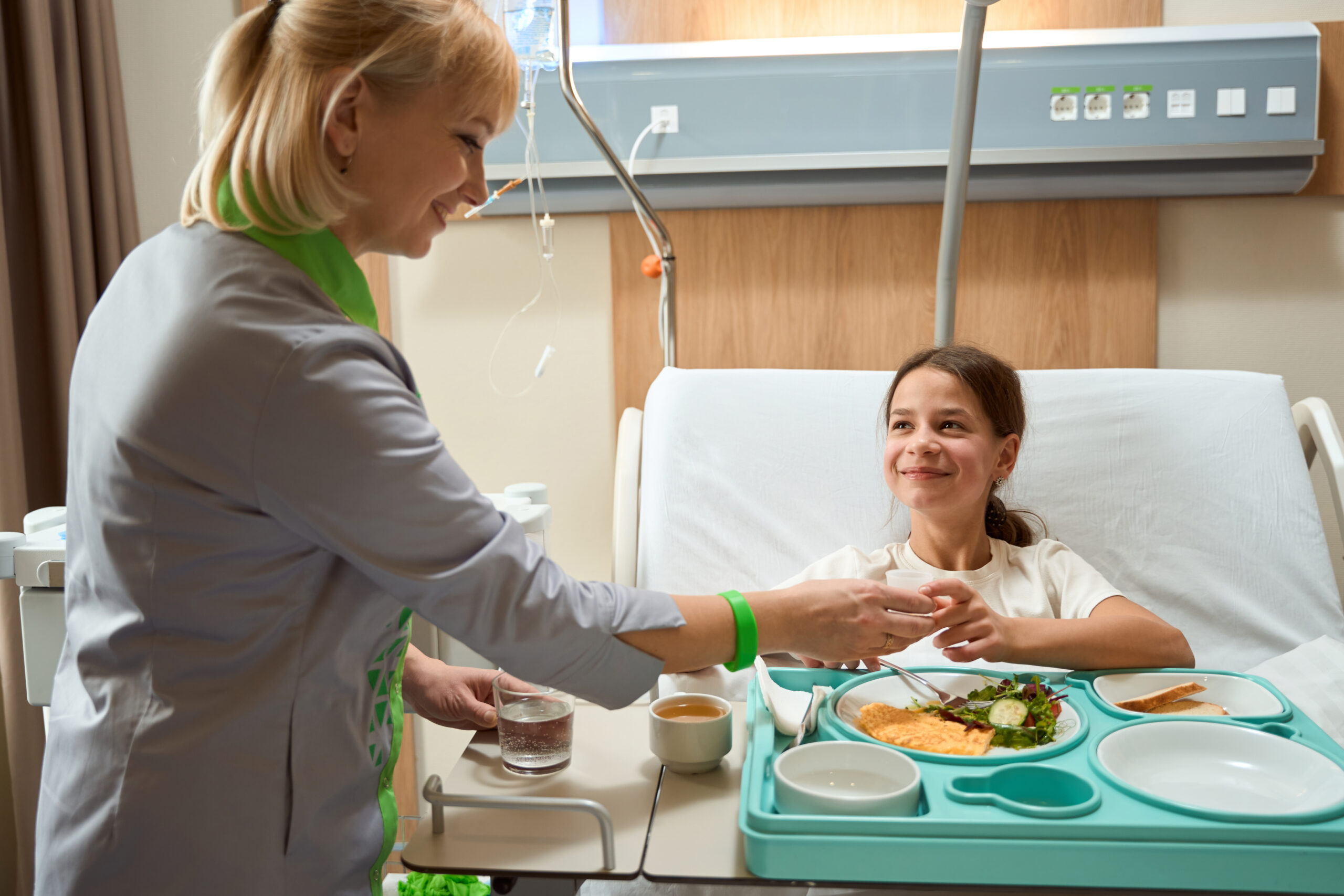 Doctor in medical clothes standing in ward and giving medicine to lady sitting on bed, eating breakfast