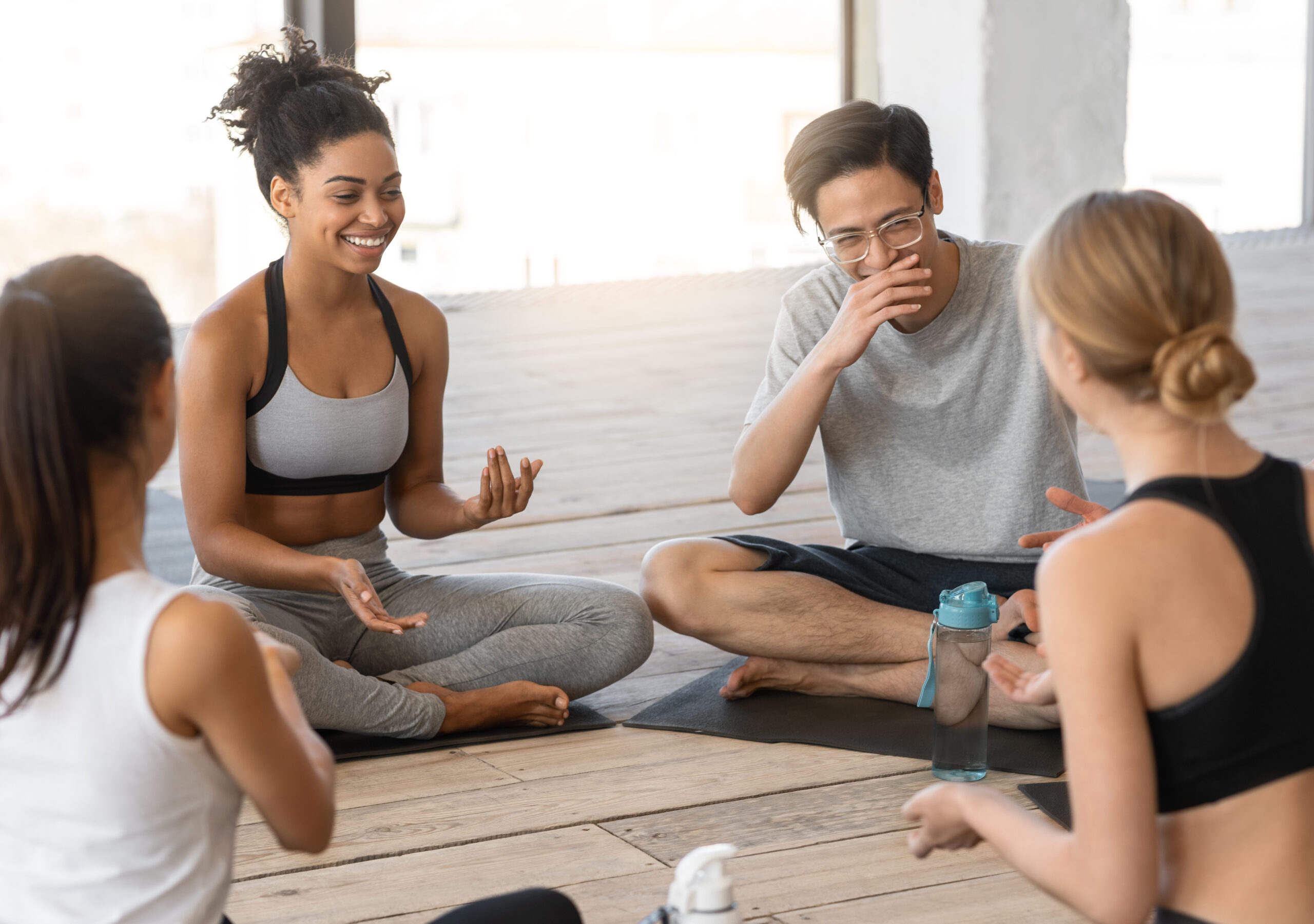 Sporty multicultural men and women sitting on mats, chatting after yoga class in modern studio, group of multiethnic people talking and smiling together after successful training, copy space